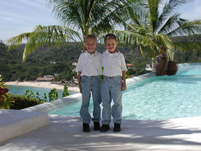 Two boys at villas entrance with palm trees, pool, beach and bay in the background.